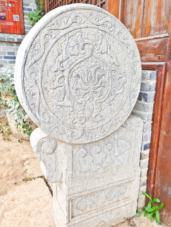 A round stone carving with intricate floral and animal patterns stands beside a wooden door and brick wall in daylight.の写真素材