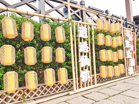 Dozens of woven yellow lanterns hang on a bamboo frame, surrounded by green foliage and Chinese characters on displayの写真素材