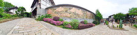 A peaceful stone path runs beside a decorative wall with mountain carvings, bordered by lush greenery and traditional architectureの写真素材