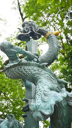 A grand, weathered bronze dragon sculpture with intricate scales twists around a tall central pillar against a green, leafy backgroundの写真素材