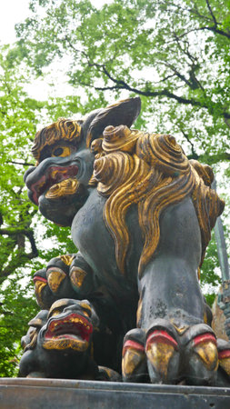 A weathered green and gold guardian lion statue is viewed from a low angle against a backdrop of treesの写真素材