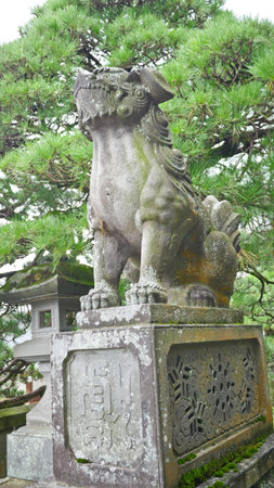 A weathered green and gold guardian lion statue is viewed from a low angle against a backdrop of treesの写真素材