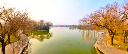Peaceful lakeside park with calm water, bare winter trees, walking paths, small bridge, and quiet scenic atmosphere under clear skyの写真素材