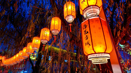 Glowing red lanterns hanging from tree branches illuminate evening pathway, creating festive...