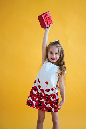 Little Caucasian girl smiling and holding red gift box on yellow background. child holding gift boxの写真素材