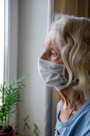Lonely old woman in a medical mask looks out the window during the lockdown. A depressed lady at home during the covid-19 pandemic.の写真素材