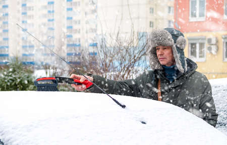 Man in a fur hat cleans snow from the roof of a car outdoors on a winter day.