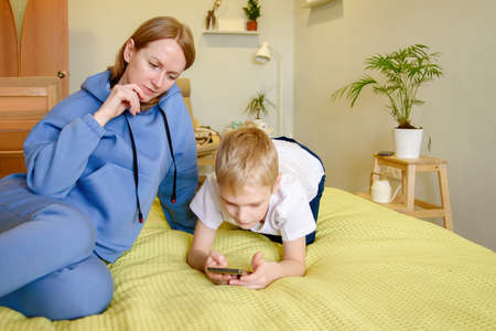 Young mother in casual clothes is lying on the bed next to her son, looking at information on the phone. Happy children with their motherの写真素材