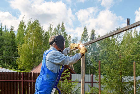 Man welder in a welding mask, construction uniform and protective gloves cooks metal on a street...
