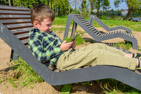 Happy little boy is sitting on a bench and playing on a mobile phone against the background of a summer park.の写真素材