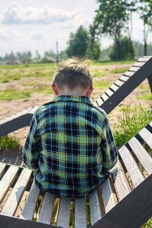 Happy little boy is sitting on a bench and playing on a mobile phone against the background of a summer parkの写真素材
