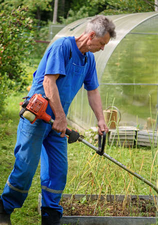 Worker with a hand trimmer in his hands mows the grass in front of the house. The trimmer is in the...