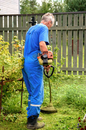 Worker with a hand trimmer in his hands mows the grass in front of the house. The trimmer is in the...