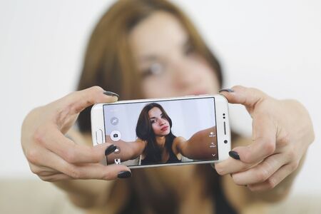 Beautiful young woman photographing herself with phone sitting in sofa