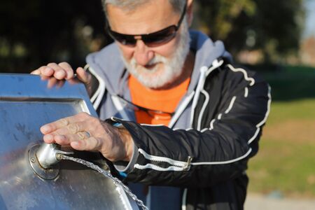 Older man with the sunglasses having his refreshment at drinking fountain in the park.の写真素材