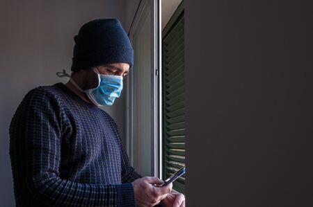 Boy with blue mask looks from a dark interior into the window light. Locked in the house by confinement. Coronavirus. Covid-19の写真素材