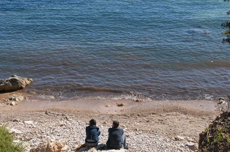 A couple with their backs to the camera and unrecognizable, sitting on a small beach in Mallorca. An image of relaxation in natureの写真素材