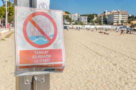No bathing sign from 21:00 to 07:00 in Spanish, Catalan and English. In the background a beach on the island of Mallorca with tourists and out of focus. New normal conceptの写真素材