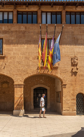 Campos, Spain; June 12 2021: Main facade of the town hall of the Majorcan town of Campos, with a tourist wearing face mask and walking under the flags of Balearic Islands, Spain and Europeのeditorial素材