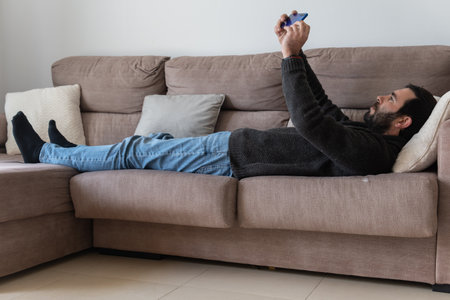 Brown caucasian man with a beard, lying on a sofa while interacting with his blue smart phoneの写真素材