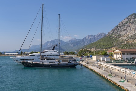 a two-masted yacht and a white boat parked at the quay berth, against the background of port buildings, green vegetation, and blue sky and mountains.の写真素材