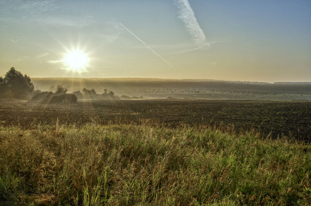 rising sun over the field, sun rays, vegetation and trees, buildings, a trail in the sky from an airplane flying at heightの写真素材