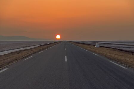 in asphalt road with road markings that goes into the distance among the desert to meet the morning rising sun.の写真素材