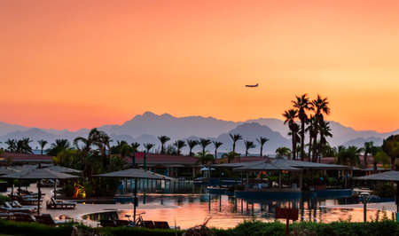 the sky painted with the colors of the sun, with mountains in the background, the silhouette of a plane flying in the sky, against the backdrop of beach umbrellas, a pool and palm treesの写真素材