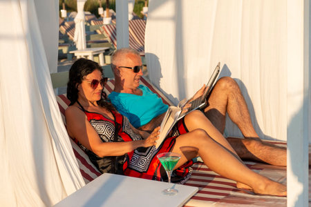 Portrait of a married couple lying on a daybed. Man working on his laptop while his wife is reading a book at the beach. Business and summer vacation - concept.の写真素材