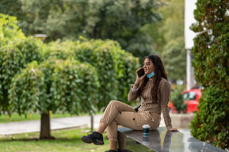 Attractive teen girl with smartphone and coffee sitting in a park and having a phone conversation....