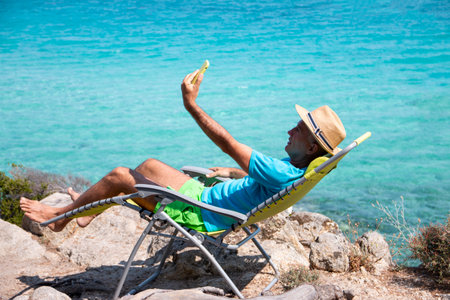 Portrait of a handsome man lying on a deckchair and and taking selfie. Happy mature man next to  the beach on Aegean sea. Summer vacation concept.の写真素材