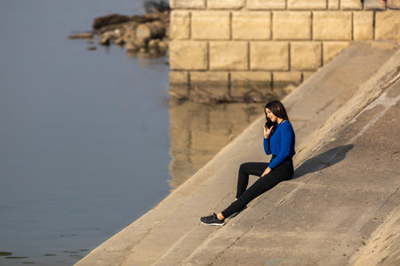 A beautiful teenager talking on her mobile phone. She is sitting on the banks of the river. Copy spaceの写真素材