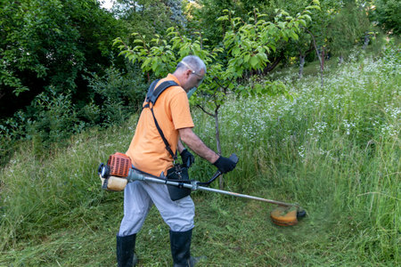A mature man in protective clothing, and gloves with a trimmer. Mowing tall grass and the weeds with...