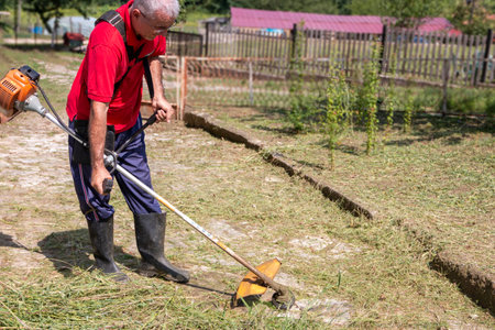 A mature man with a trimmer cuts grass and weeds with a trimmer on a paved path.の写真素材