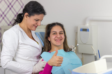 Satisfied  patient. Teenager girl showing thumb up and smiles  while sitting  at dentist's  chair after dentalの写真素材