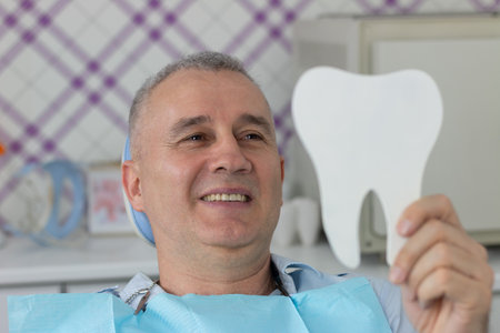 A middle aged man is sitting in the dental chair and using a mirror to see the result of the stomatology work which was doneの写真素材