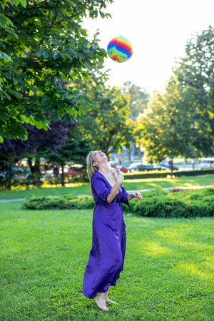 Attractive Woman with Long Hair and Violet Dress is Enjoying in Playing in Public Park with Colorful Ball.の写真素材
