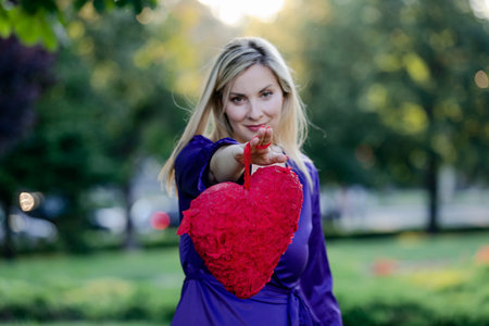 Attractive Woman with Long Hair and Violet Dress is Walking in Public Park and Holding a Red Heart.の写真素材
