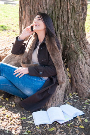 Beautiful teenage girl reads the book under the huge oak tree on sunny autumn day and using a smart phone.の写真素材