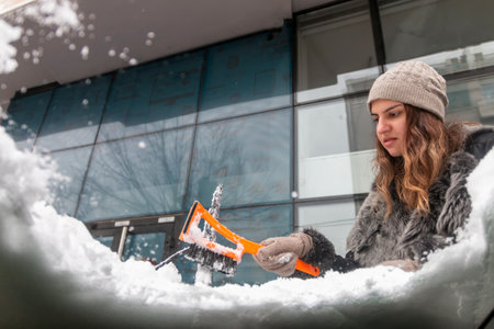 Young  woman using a snow brush to remove snow from her car's windshield. Concept of winter weather, car maintenance, and being prepared for snowy conditions. Transportation conceptの写真素材