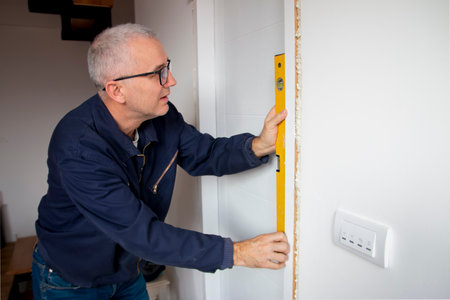 Male repairman in a working suite and with blue hat measuring door frame with a spirit level.