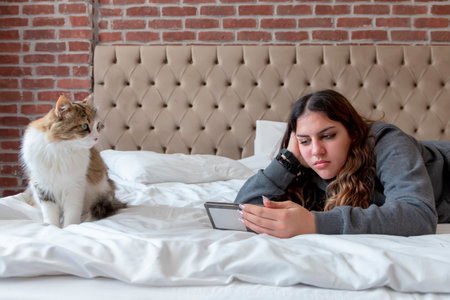 Young woman laying on the bed watching movie on her phone, while her cat is watching.の写真素材