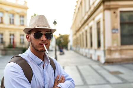 Handsome Well Dressed Young Man with Hat, Sunglasses and Backpack Standing in the City Center and Smoking a Cigaretteの写真素材