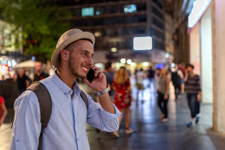 Handsome Young Man with Hat and Backpack Standing in the City Street and Talking over his Mobile Phoneの写真素材
