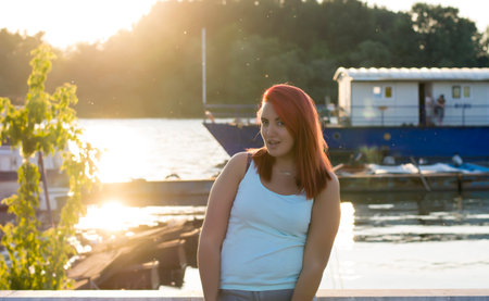 Beautiful long haired redhead is posing near the river bank during sunsetの写真素材