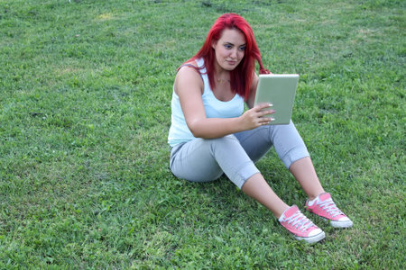 Attractive redhead woman sitting on the grass in park and using her digital tabletの写真素材