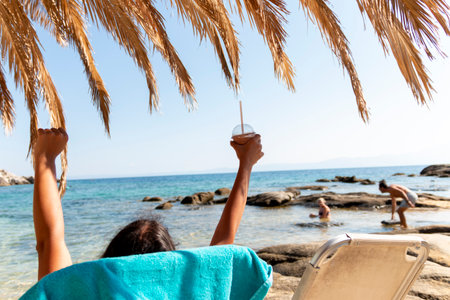 A young woman sits on a comfortable beach chair, enjoying a refreshing cocktail as she gazes out at the crystal-clear ocean.の写真素材