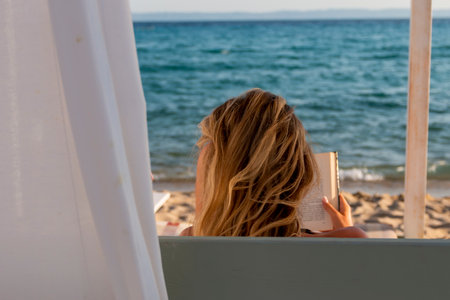 Young blonde woman reading a book and relaxing on a daybed at the beachの写真素材