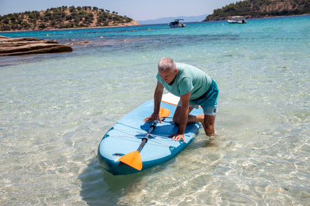 Sup surfing. Beautiful view of the sea with a mature man kneeling on a board with paddle in the water. Standup paddleboarding in summerの写真素材