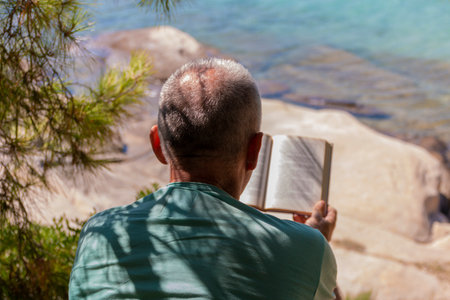Mature man lying on a sunbed and reading a book. Happy mature man next to the beach on Aegean sea with beautiful view. Summer vacation concept.の写真素材
